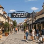 Shoppers stroll through La Vallée Village outlet at Val d’Europe near Disneyland Paris, surrounded by designer boutiques like Burberry and Moncler on a sunny day.