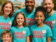 A diverse group of smiling adults and children wearing matching “Birthday Squad” t-shirts and badges, posing together at a colourful theme park entrance. Perfect for celebrating special occasions with friends and family in 2025.