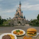 Plates of theme park food—steak and chips, buffet meal, burger with fries, Mickey-shaped waffle and macaron—in front of a fairytale castle, representing table service, buffet, and quick service dining options.