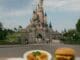 Plates of theme park food—steak and chips, buffet meal, burger with fries, Mickey-shaped waffle and macaron—in front of a fairytale castle, representing table service, buffet, and quick service dining options.